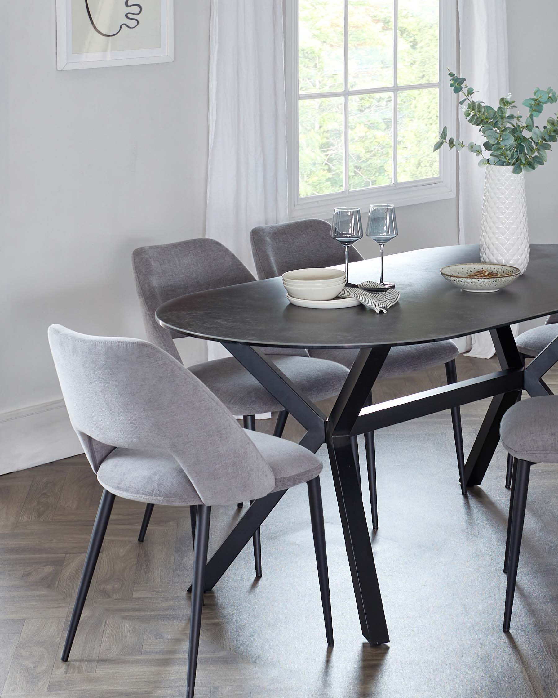 A modern dining set featuring a round, black table with a matte finish and four angled legs, accompanied by four upholstered chairs in light grey fabric with curved backs and slender black metal legs, displayed in a bright room with sheer curtains and wood flooring.