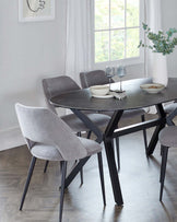 A modern dining set featuring a round table with a dark, smooth surface and angular black metal legs. Accompanied by four elegant upholstered chairs in light grey fabric, boasting sleek lines and slender black metal legs. The set is positioned on a hardwood floor, complemented by a minimalist decor with a large window draped with white curtains in the background.