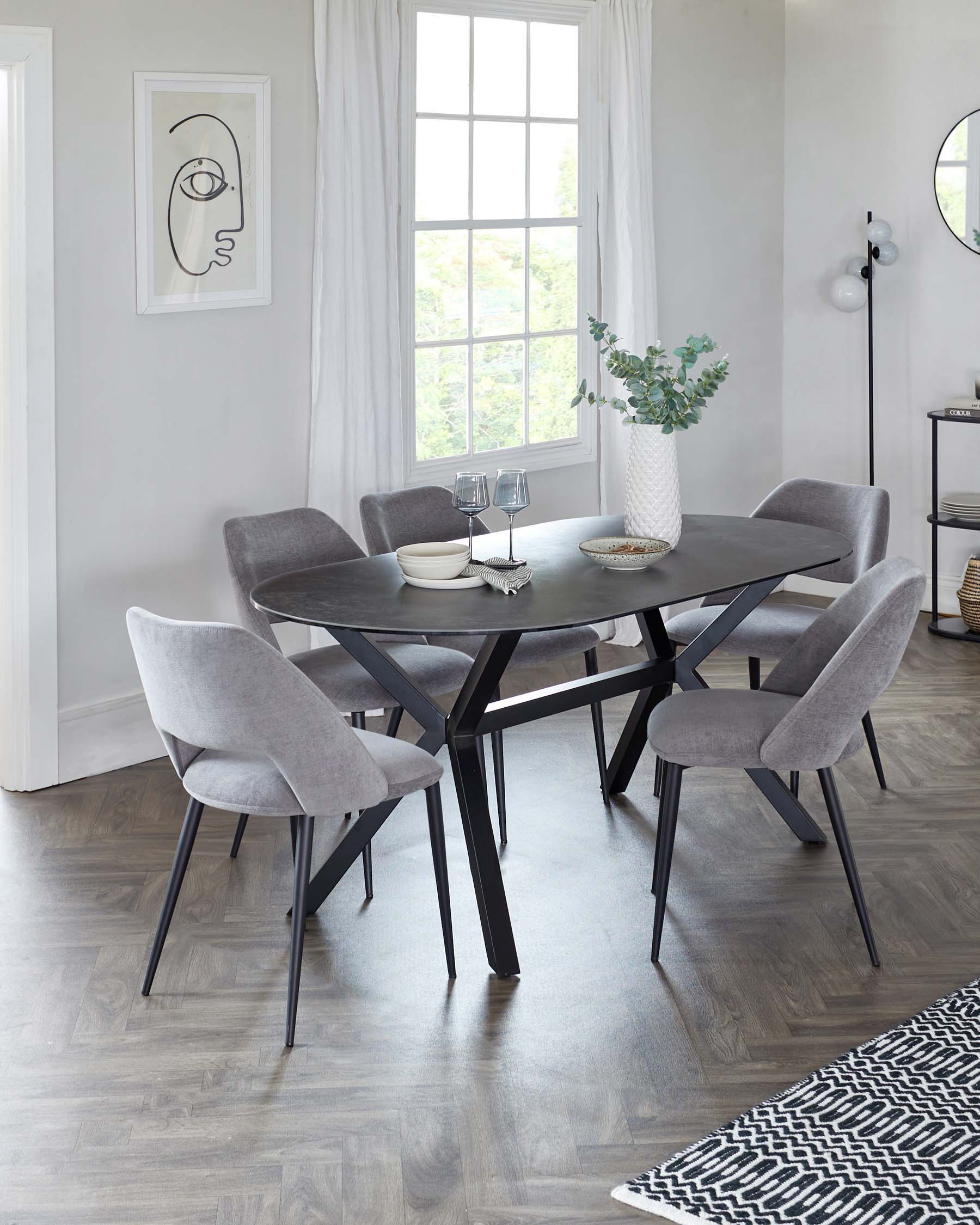 A modern dining set with a black, round table featuring angular legs and a matte finish. Surrounded by six grey, upholstered chairs with a sleek design and black legs, set on a light wooden floor. The table is accented with a textured white vase containing greenery, and the setting is lit by natural light from a window.