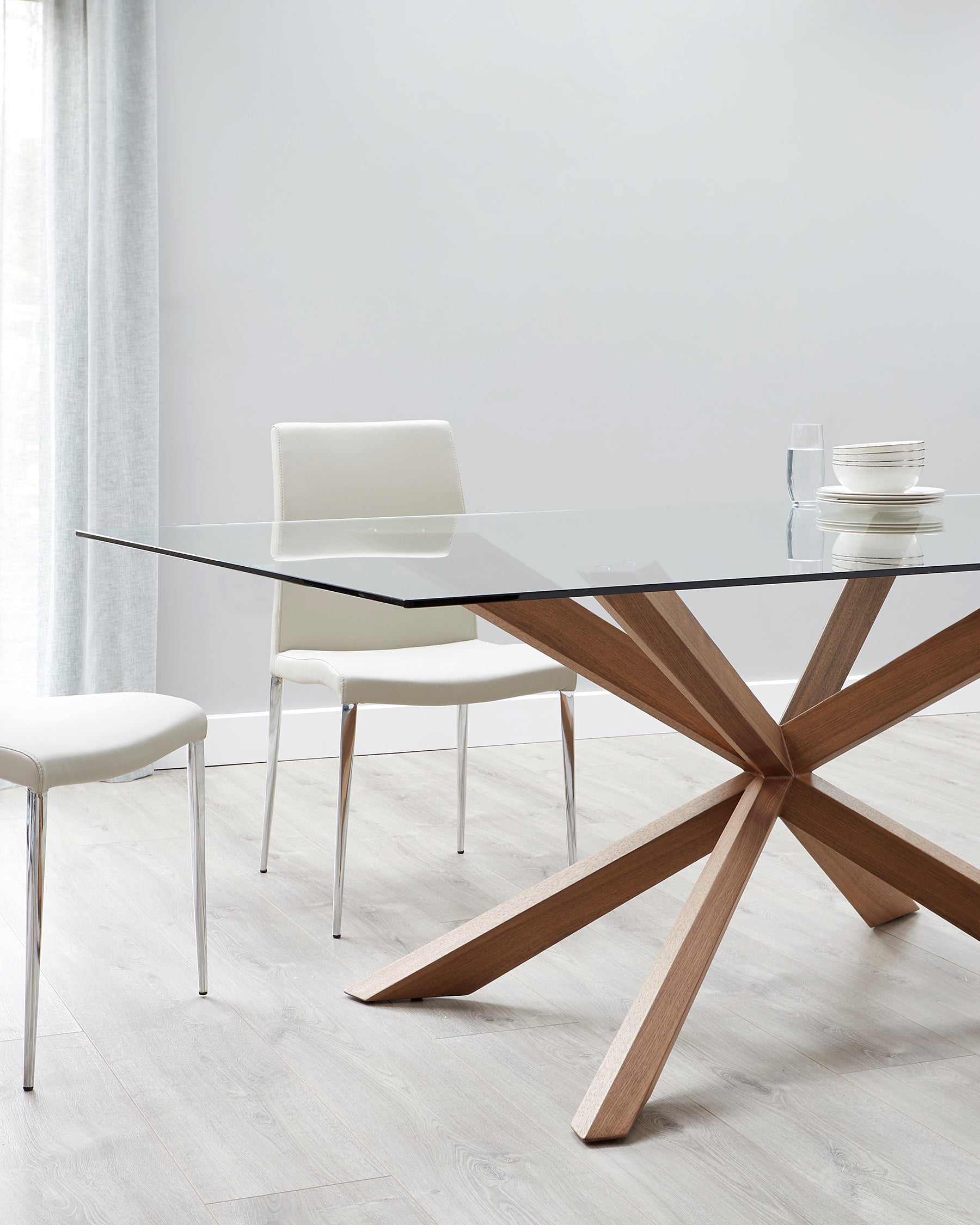 Modern dining room furniture featuring a rectangular glass-top table with a distinctive wooden starburst-style base and three sleek chairs with white upholstery and chrome legs, complemented by a simple set of white dishes on the table.