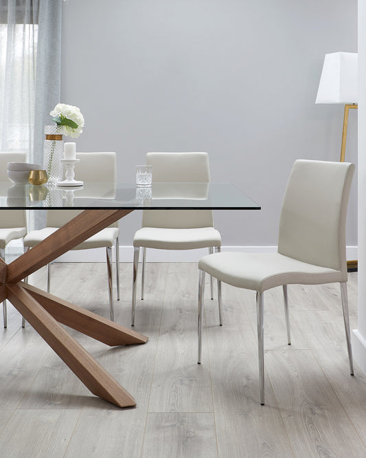 Modern dining room featuring a rectangular glass tabletop with a unique crossed wooden base, paired with six white upholstered chairs with sleek metal legs. A floor-standing lamp with a white shade complements the ensemble.