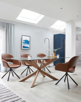 A modern dining room set comprising a rectangular glass tabletop resting on a sculptural wooden cross base and four matching chairs with brown leather upholstery and black metal legs.