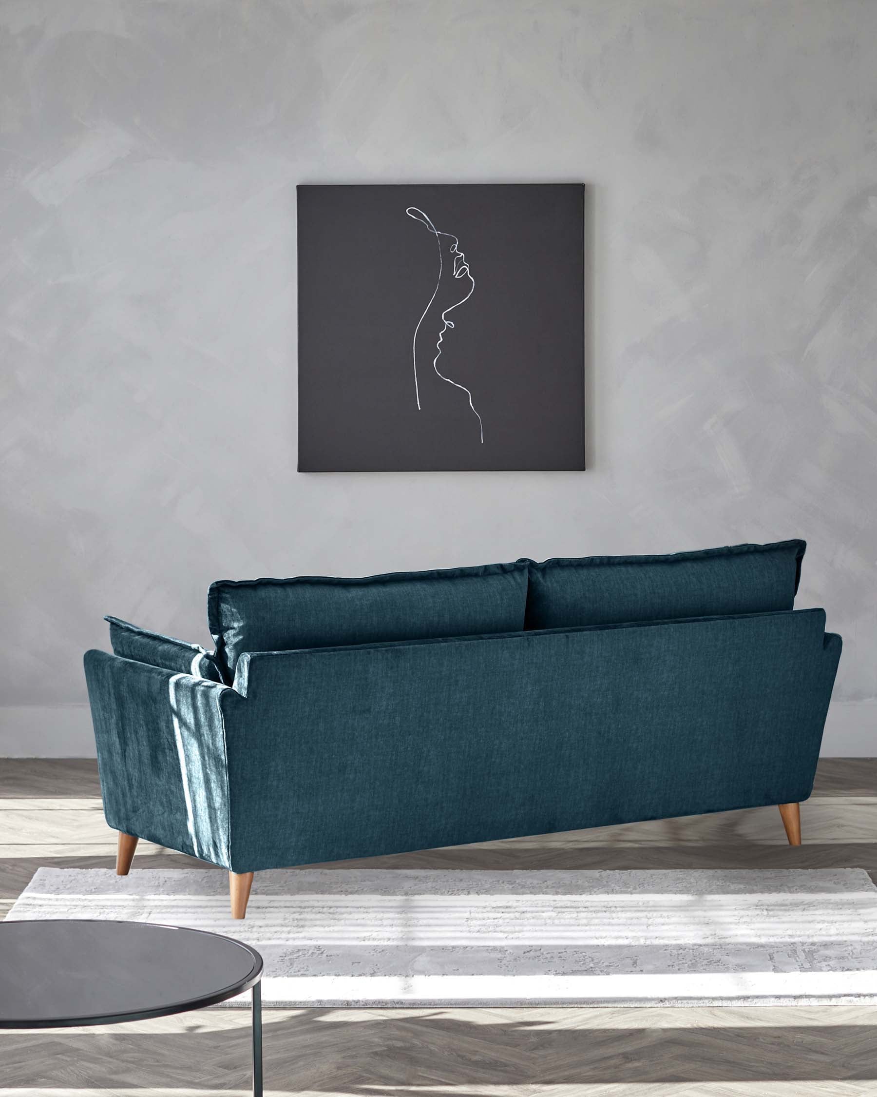 Elegant teal velvet three-seater sofa with a tufted backrest and angled wooden legs. In front, a minimalist round side table with a black top and thin metal legs. The room is completed with a textured grey wall and a white-grey patterned area rug.