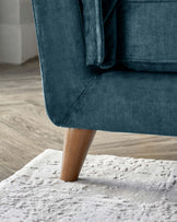 Close-up of a modern sofa corner with a blue fabric upholstery and a sleek tapered wooden leg, standing on a plush white area rug with a textured pattern, against a backdrop of light wood flooring and a white wall.