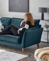 A modern teal fabric three-seater sofa with plush cushions and angled wooden legs. There's also a white and grey area rug partially visible at the bottom, and a side table with a black lamp and decorative items in the background.