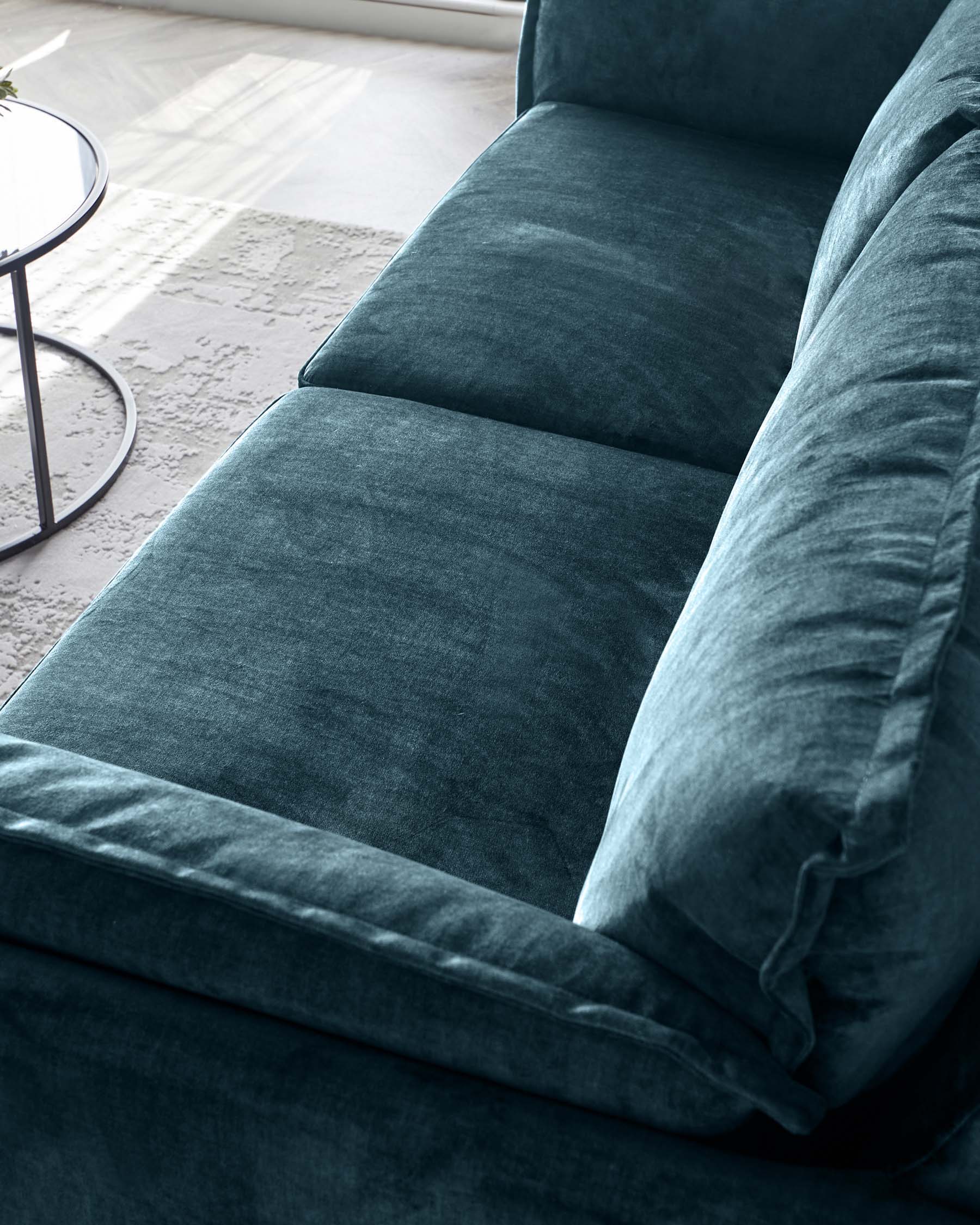 A modern deep teal velvet sofa, featuring clean lines, rectangular cushions and an armrest on one side. A plush back cushion and a sleek metal and glass side table are partially visible alongside. The sofa is set against a light, airy room with a white curtain and a textured area rug.