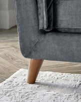 Close-up view of a modern sofa with grey upholstery and a tapered natural wood leg, standing on a plush white textured area rug on a wooden floor.