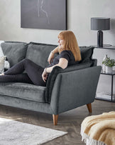 A plush, modern grey fabric sofa with three seat cushions, flared armrests, and tapered wooden legs. Nearby, a sleek black metal table lamp with a matching circular shade sits atop a minimalist black end table, accompanied by decorative items including a white vase with greenery. The setting is completed with a textured white area rug partially under the sofa.