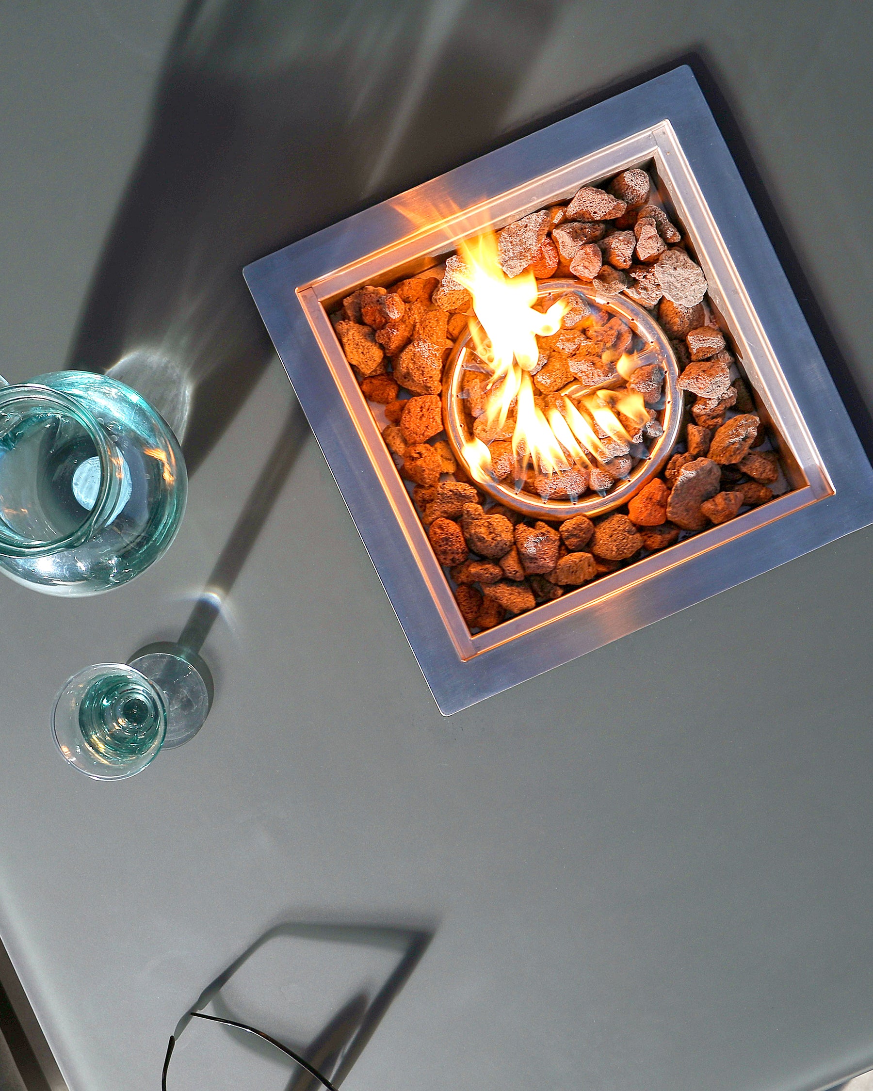 Modern outdoor square fire pit table with a built-in central gas fire surrounded by natural lava rocks, and two transparent wine glasses placed on one corner of the table.