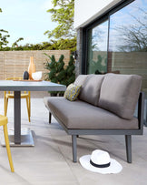 Outdoor furniture set featuring a modern grey two-seater sofa with clean lines and dark frame, complemented by a sleek contemporary dining table with a grey surface and vibrant yellow legs. A contrasting decorative pillow with intricate patterns adds a touch of colour to the setting.