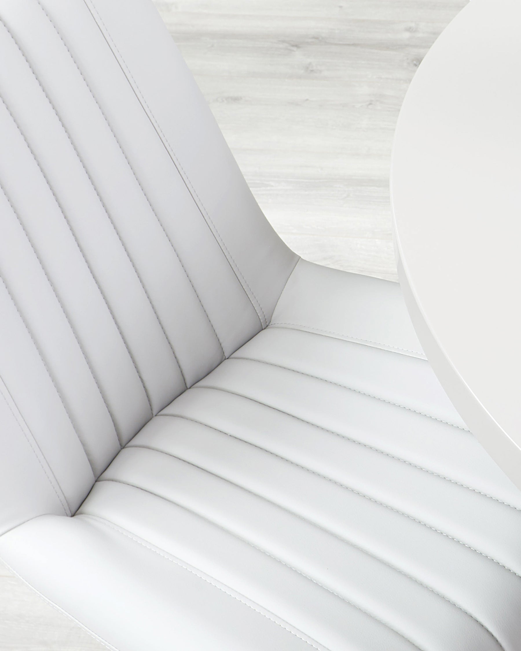 Modern white faux leather chair with vertical stitching details and a high backrest, accompanied by a portion of a round white table, against a light wood grain flooring background.