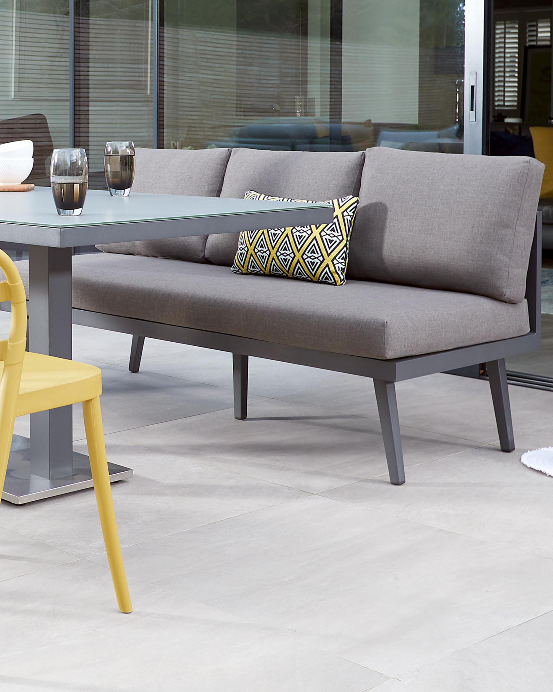 Modern outdoor furniture featuring a sleek, grey upholstered corner sofa with clean lines and a matching rectangular glass-top coffee table with a metal frame. A vibrant yellow chair with a contemporary design adds a pop of colour to the setting.