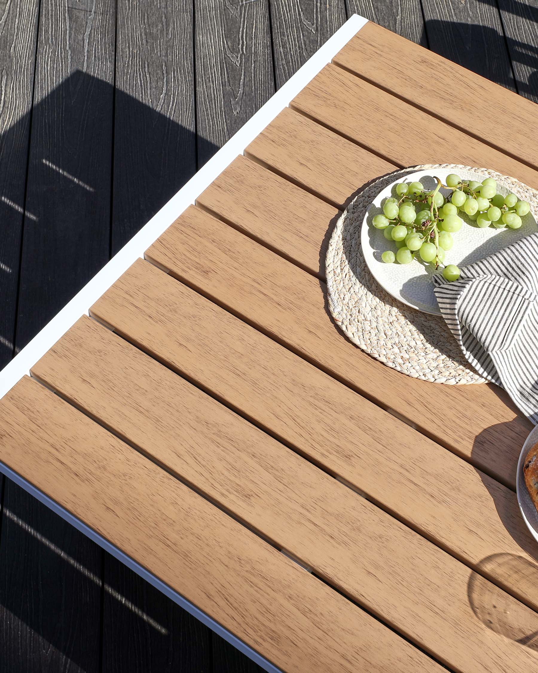 A modern outdoor hardwood dining table with parallel plank design and clean white edging, set atop a dark wood deck, partially covered by a textured circular placemat with a cluster of green grapes and a striped cloth napkin.