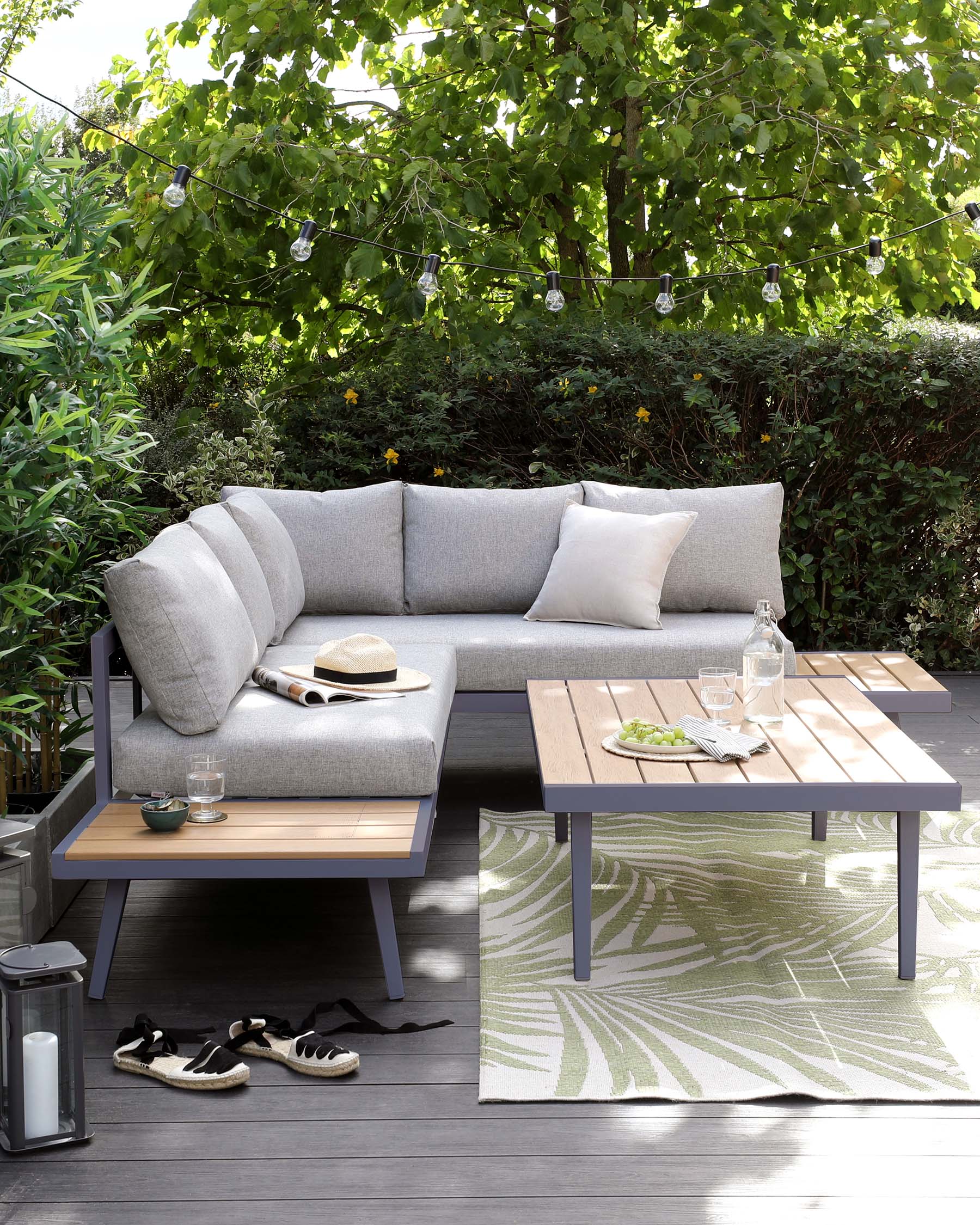 Outdoor furniture set featuring a modern grey sectional sofa with plush cushions and throw pillows, paired with a sleek purple metal-framed coffee table with a natural wood tabletop. The set is accented by a green and white patterned outdoor rug, all staged on a wooden deck with green foliage in the background.