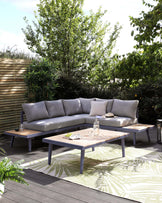 Outdoor sectional corner sofa with light grey cushions and two matching wooden coffee tables with grey metal legs, displayed on a wooden deck with greenery in the background.