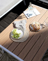 Modern outdoor wooden table with a white frame, accompanied by a partial view of a coordinating bench with beige upholstery, set on a patterned outdoor rug.