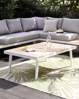 Outdoor furniture set featuring a white aluminium frame sectional sofa with light grey cushions and a matching low-profile white aluminium coffee table with light wooden slats on top. The set is arranged on a patterned outdoor rug with palm leaf design.