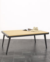 A modern rectangular coffee table with a ribbed light wooden tabletop and slender, straight metal legs in a matte black finish. The table is adorned with a casual straw hat and a pair of sunglasses resting atop an open magazine. The furniture piece is set against a neutral background with grey floor tiles.