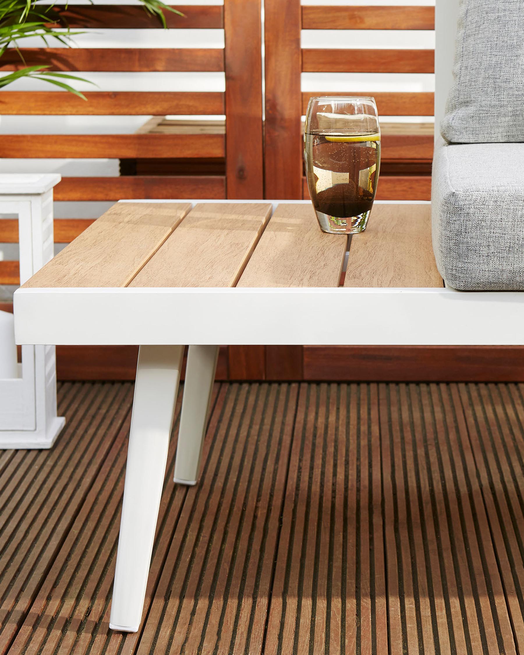 Modern outdoor patio furniture featuring a white-framed coffee table with natural wooden top planks, complemented by a part of a grey cushioned sofa arm visible on the right. A clear glass with a beverage and lemon slice rests atop the table, with a wooden slatted backdrop adding warmth to the cosy, inviting setup.