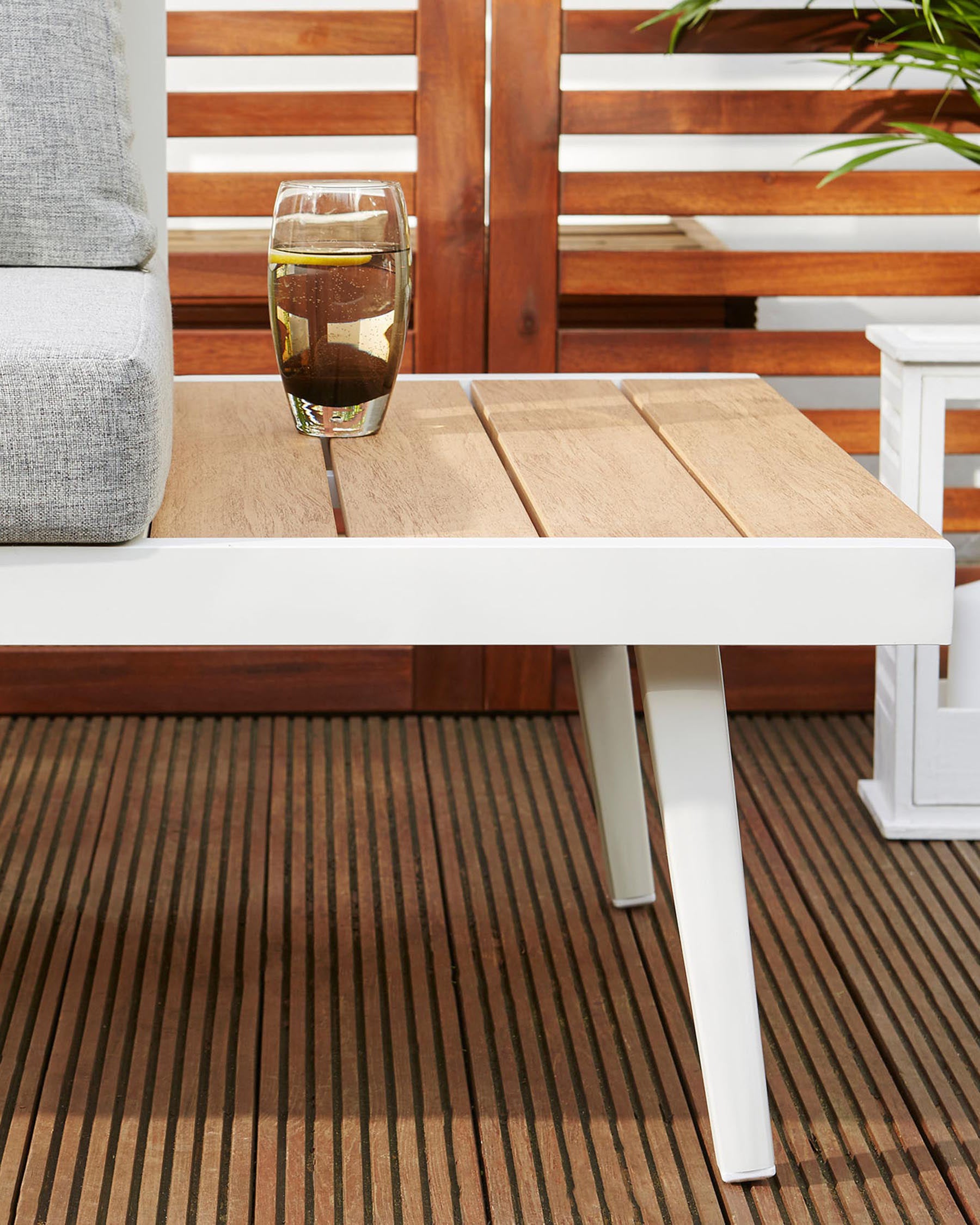 Contemporary outdoor furniture setting featuring a white-framed low coffee table with natural wood-finished tabletop, complemented by a grey cushioned corner seat with white base. A glass of drink rests on the table, set against a backdrop of a wooden panel fence and a striped brown deck floor.