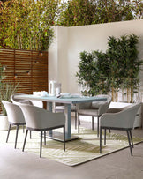 Modern outdoor dining furniture set featuring a rectangular, light blue table with a sleek finish and solid legs, paired with six upholstered armchairs in a complementary grey fabric that covers both the seats and the backrests. The chairs showcase slender, dark-toned metal legs, providing a contemporary yet comfortable dining experience. The set is arranged on a patterned area rug with green leaf motifs, adding a touch of nature-inspired design to the space.