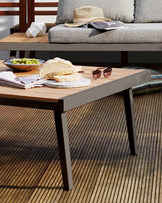 Modern outdoor lounge setting featuring a sleek, rectangular wooden coffee table with a flush top and slender, angled, dark metal legs, accompanied by a light grey cushioned sofa with clean lines and a textured fabric. The setting is accented with a straw hat and reading materials on the sofa, and a casual food arrangement on the coffee table, highlighting a relaxed, contemporary lifestyle.