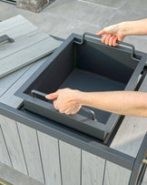 A modern outdoor furniture set featuring a grey wooden table with a black removable tray being handled by a person, revealing outdoor storage space underneath. The functional design offers versatility for dining and storage solutions on a patio or deck setting.