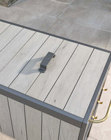 Modern outdoor patio table with a sleek design, featuring a light grey wooden plank top with a matte finish, framed by a dark grey metal edge. The table showcases unique metal handles on each side, highlighted with a golden finish, which adds a touch of elegance to the minimalist aesthetic. The table is positioned on a grey tiled floor, suggestive of a contemporary outdoor or indoor setting.