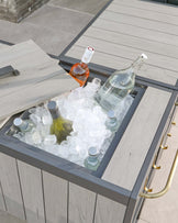 Modern outdoor patio cooler table featuring a grey wood finish, framed with a dark metal structure, and accented with gold-toned handles. The table includes a built-in ice bucket compartment filled with ice and several bottles, with a sliding wood cover partially covering it.