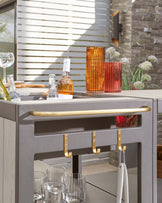 Modern outdoor grey bar cart with golden handles and towel racks, featuring two levels – a lower shelf for storing glasses and an upper drawer for utensils.