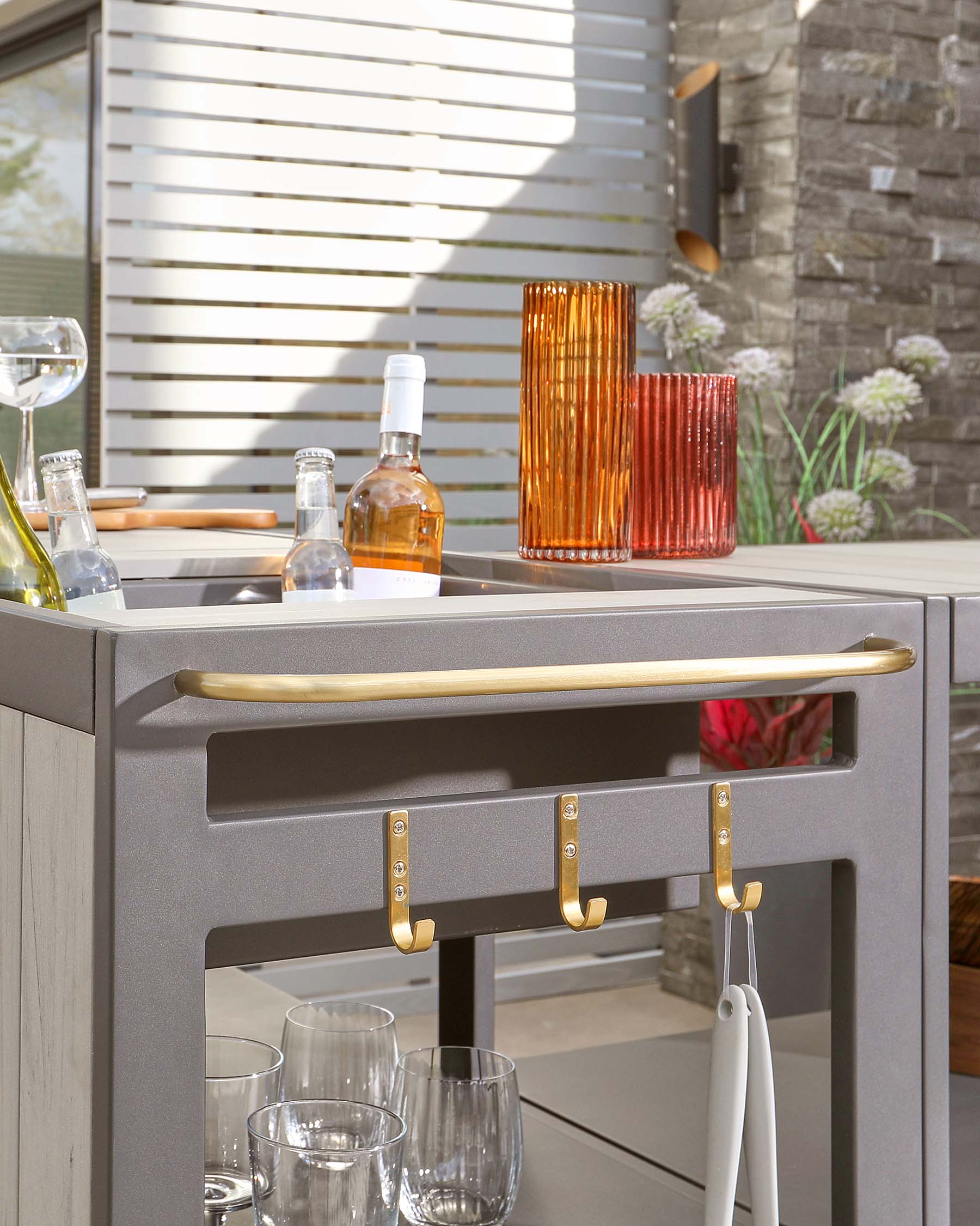 Modern grey outdoor bar cart with golden handles and accents, featuring two storage shelves and a set of hanging hooks for utensils or towels.