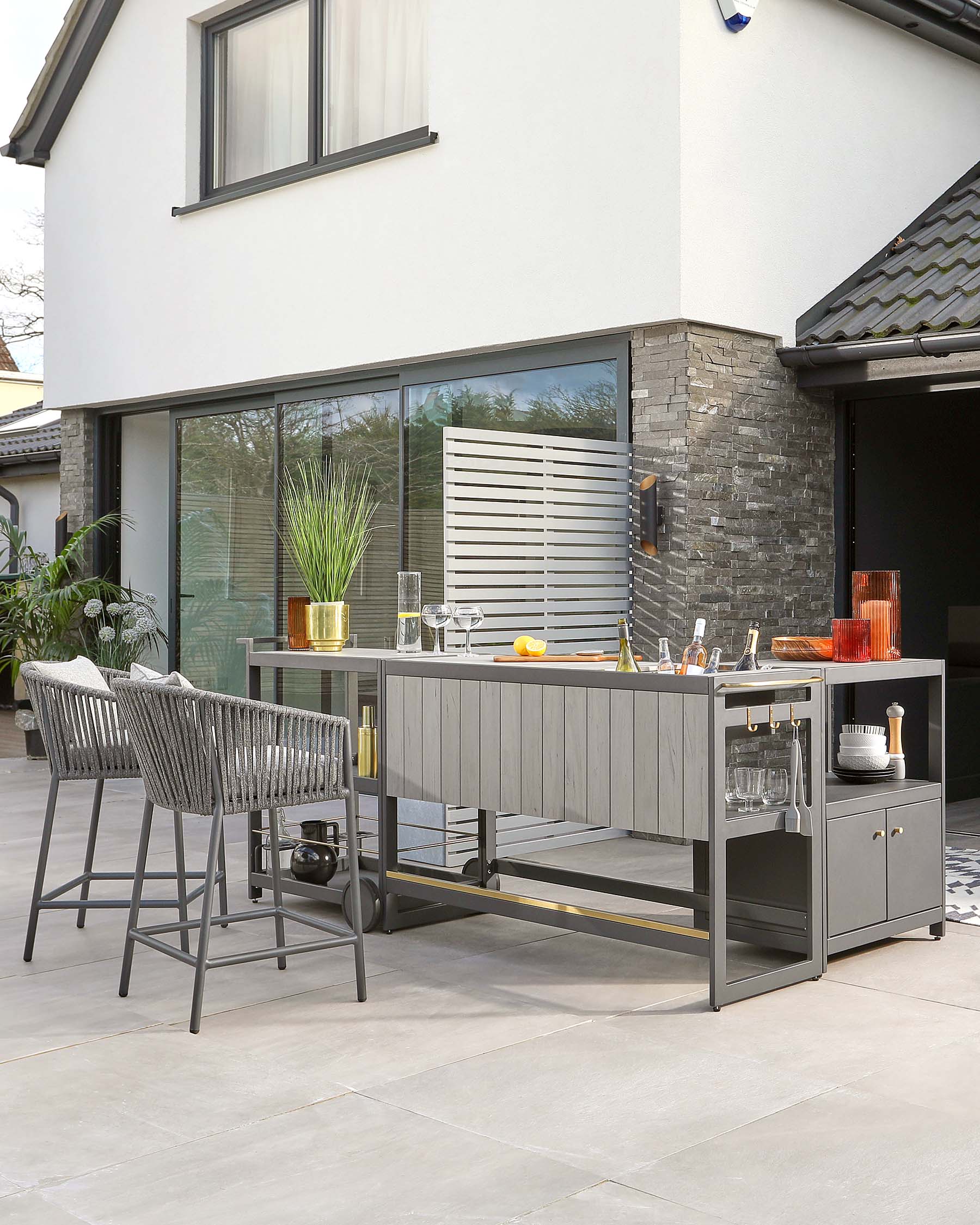 Contemporary outdoor bar set featuring a grey high-top table with clean lines, integrated shelving on one side, and space for bar seating. Accompanied by two stylish bar chairs with woven backrests and seats, and slender, angled metal legs that complement the table's modern aesthetic.