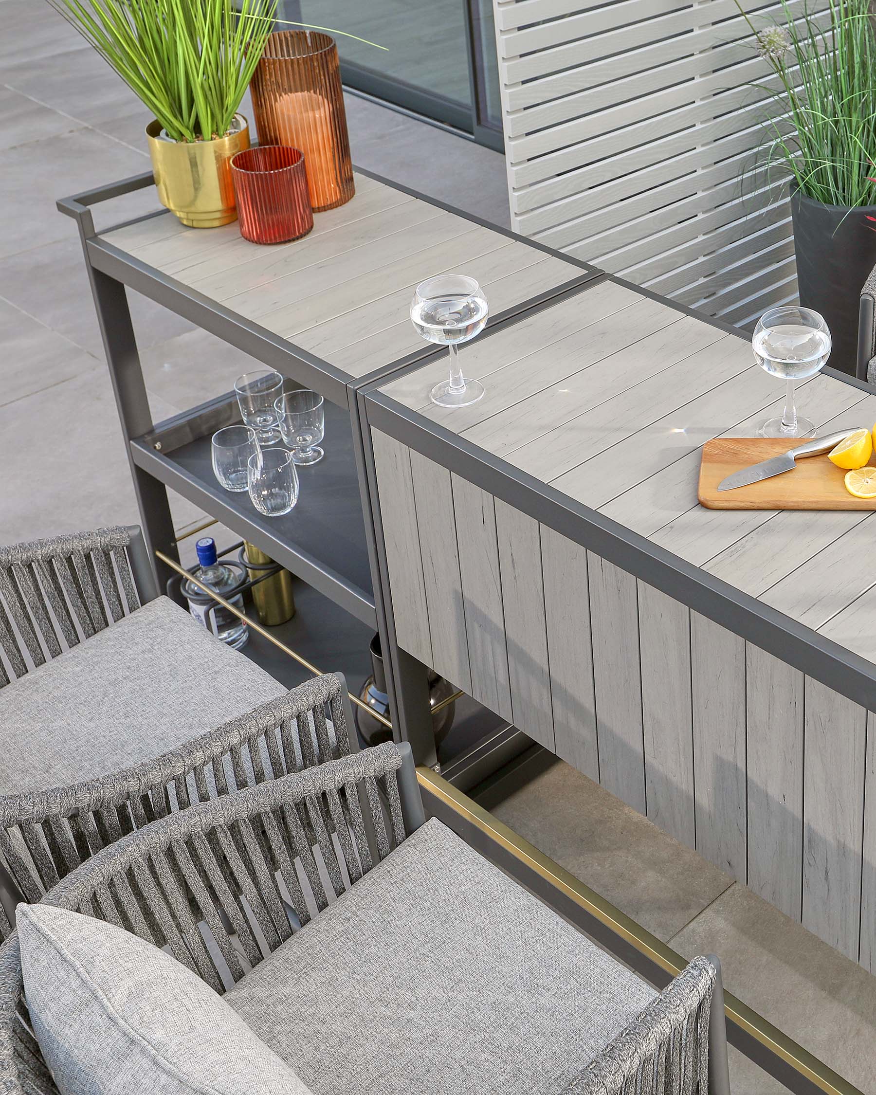 Modern outdoor furniture including a grey wood-textured aluminium dining table with a central storage rack and matching chairs featuring dark grey woven fabric and metallic frames. Decor includes glassware and potted plants.