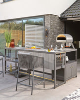 Contemporary outdoor kitchen furniture set featuring a dark grey modular island with integrated storage and workspace, complemented by a high bar chair with grey textured upholstery. An elegant bar trolley with bottles and glasses is positioned to the side. The set is designed for sophisticated al fresco cooking and entertaining.