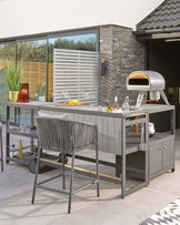 Modern outdoor kitchen station with a matte grey finish and stainless steel accents, featuring storage cabinets, a countertop, and a space for a pizza oven. Accompanied by a matching high bar stool with a woven backrest and a metallic frame. A rolling bar cart with glassware storage is also visible in the background.