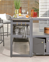 Modern outdoor grey bar cart with a stainless steel top, featuring storage shelves, a drawer, and space for glassware. Accented with gold-tone handles and casters for mobility.