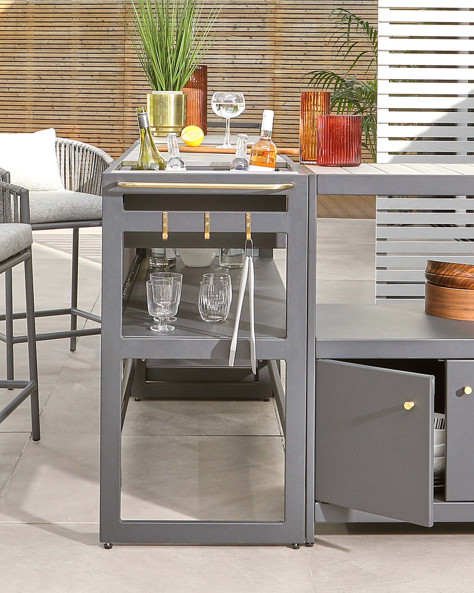 Modern outdoor grey bar cart with a stainless steel top, featuring storage shelves, a drawer, and space for glassware. Accented with gold-tone handles and casters for mobility.