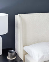 A modern off-white upholstered headboard with a plush texture paired with a soft white pillow on a matching bed frame. A stylish black side table with a textured black ceramic mug resting on a coaster complements the clean, contemporary look, and a sleek white table lamp with a rounded base and cylindrical shade adds to the minimalist aesthetic. The furniture is set against a dark wall, highlighting the contrast and creating a sophisticated bedroom ambiance.