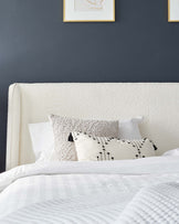 A modern bedroom showcasing a plush, cream-colored upholstered headboard paired with a neatly made bed clothed in white linens with a subtle striped pattern. A variety of textured decorative pillows, including one with a pattern of black embroidered lines and tufts, accents the bed setup. The wall behind is painted a deep blue, providing a stark contrast to the light tones of the bedding, and is adorned with two framed abstract art pieces with gold trim.