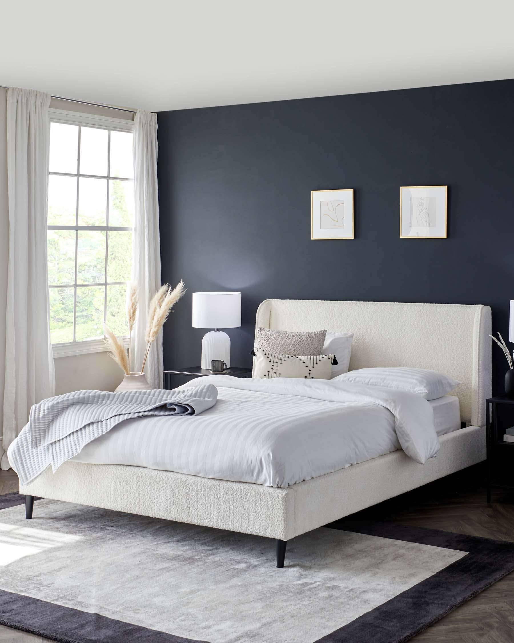 Elegant modern bedroom showcasing a large upholstered bed with a textured cream-colored fabric headboard and frame, flanked by two black nightstands with smooth finishes. The bed is accessorized with crisp white bedding accented by a decorative throw and pillows. A white table lamp with a round base sits atop one nightstand, complementing the room's minimalist aesthetic. The entire setting is anchored by a two-tone grey area rug, adding a layer of warmth and sophistication to the space.