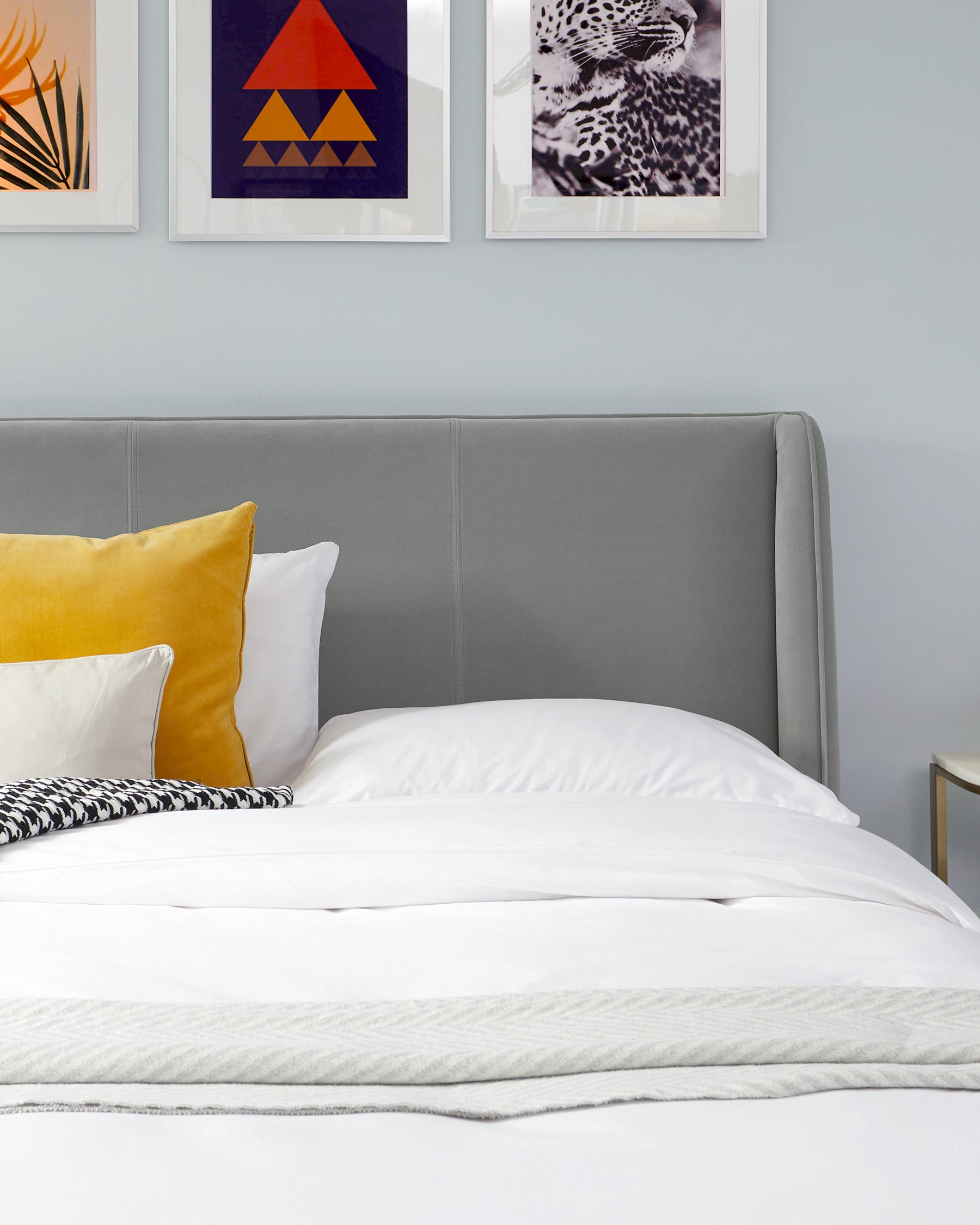 Contemporary upholstered grey headboard with clean lines and a padded, stitched panel design, accompanied by a white bedspread, complemented by various pillows including a yellow velvet one, a white one, and another with zigzag patterns.