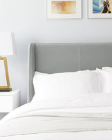 Elegant grey upholstered headboard with a simple, clean design paired with a minimalist white nightstand featuring a single drawer. A gold-coloured lamp with a white lampshade complements the modern and sleek bedroom furniture setup.