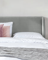 Elegant modern bed with a tall, gently curved, grey upholstered headboard and visible stitching details, complemented by white bedding and a decorative textured throw at the foot of the bed. One visible pink and one white pillow add a soft colour accent.