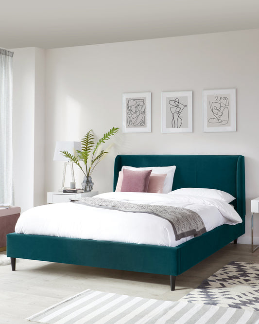 Elegant emerald green upholstered bed with a high headboard and dark wooden legs, flanked by a white modern nightstand with a lamp and decorative plant. A grey and white striped area rug lies at the foot of the bed.