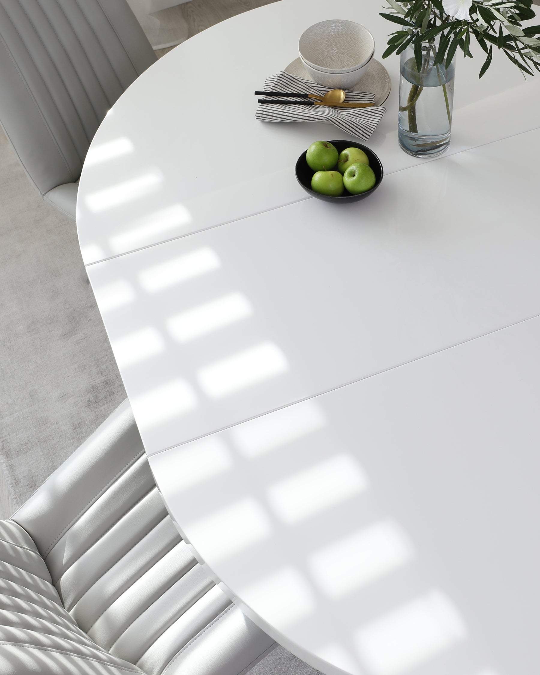 A high-gloss white round dining table with a seamless surface, accompanied by sleek, mid-back chairs upholstered in light grey with vertical channel stitching. The table is adorned with a black bowl containing green apples, a striped napkin with cutlery, a textured grey bowl, and a clear vase with white flowers. The sunlight creates a pattern of bright rectangles on the tabletop's surface.