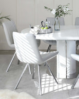 A modern dining set featuring a white, round table with a glossy finish and sleek, metallic legs paired with comfortable white leather chairs with vertical stitching detail and slim chrome-finished legs. The scene is complemented with a woven tray, a glass vase with white flowers, and a minimalistic coffee maker placed on the table, enhancing the clean and contemporary aesthetic.