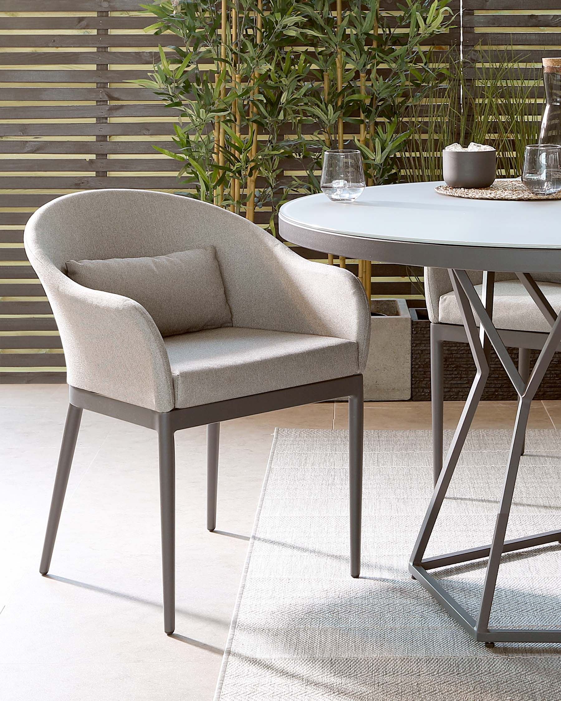 Modern outdoor furniture set featuring a curved-back light grey fabric chair with metal legs, and a round white-top table with an angular metal base, arranged on a textured light beige rug, with a backdrop of green plants and a slatted wood screen.