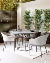 A contemporary outdoor dining set consisting of a round, grey tabletop supported by a geometric metallic base and four upholstered armchairs with sleek, metal legs. The chairs feature a soft, curved backrest and comfortable seating, in a light, neutral fabric that complements the modern design. The set is arranged on a textured white outdoor rug, accentuating its elegant and sophisticated silhouette.