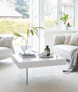 Elegant white tufted sofa with matching armchair, complemented by a modern white coffee table with transparent glass legs. The setting is styled with minimalistic décor and plush carpeting, enhancing a light and airy room ambiance.