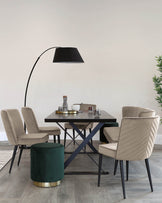 Modern dining set featuring a square black table with a distinctive blue x-shaped metal base, complemented by three beige upholstered chairs with vertical stitching and slim black metal legs. A dark green velvet ottoman with a gold metal base sits next to the table. A sleek, large black floor lamp with an arched neck looms overhead.